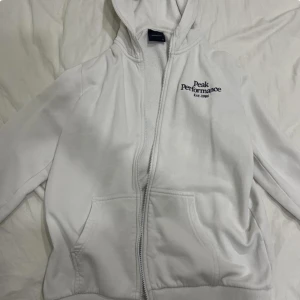 Vit hoodie från Peak Performance - Säljer en vit hoodie från Peak Performance med dragkedja och broderad logga på bröstet. Den har en bekväm passform och är perfekt för kyligare dagar. Huvan ger extra värme och stil. STORLEK: XS