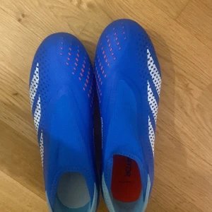 Blå fotbollsskor från Adidas - Snygga blå fotbollsskor från Adidas med vita och röda detaljer. Skorna har en strömlinjeformad design och är perfekta för fotbollsplanen. De har en mönstrad yta för bättre grepp och kontroll. Pris kan snackas privat.