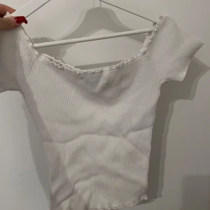 Vit ribbad topp från new yorker  - Säljer en söt vit ribbad topp från new yorker. Den har en offshoulder-design med korta ärmar och en fin volangdetalj vid halsringningen. Perfekt för en stilren och trendig look. Använd 5 gånger för 3 år sedan !!