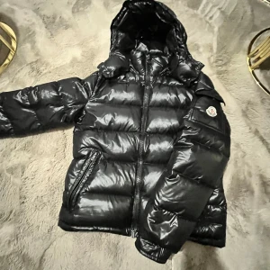 Svart dunjacka från Moncler - Säljer en snygg svart dunjacka från Moncler med glansig finish. Jackan har en huva och dragkedja framtill samt Moncler-logga på ärmen. Perfekt för kyliga dagar.