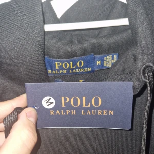 Svart hoodie från Polo Ralph Lauren - Säljer en stilren svart hoodie från Polo Ralph Lauren med en liten röd logga på bröstet. Tröjan har en praktisk känguruficka och justerbar huva med snören. Perfekt för en avslappnad look.