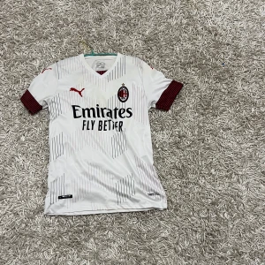 AC Milan fotbollströja - Snygg vit AC Milan fotbollströja från Puma med röda detaljer och svart text. Tröjan har korta ärmar och klubbens emblem på bröstet. Perfekt för fans som vill visa sitt stöd.