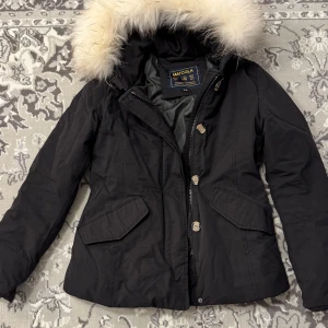 Svart jacka med vit pälskrage från Matogla - Snygg svart vinterjacka från Matogla med riktig vit pälskrage. Jackan har knappar och dragkedja framtill samt två jackfickor, ingen innerficka. Huvan kan inte dras av men pälsen är avtagbar. Material: 80% bomull, 20% polyester. Perfekt för kyliga dagar.