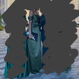 Grön axelbandslös balklänning - Elegant oliv grön långklänning i satin med offshoulder-design och draperad överdel. Klänningen har vackra detaljer med glittrande dekorationer vid axlarna och en figursydd passform som framhäver midjan. Går att justera tyget vid brösten antingen kan man visa korsetten lite eller dra upp tyget så det blir en vanlig fin överdel. (På bild 1 är tyget uppe och på bild 2 syns korsetten) använt 2 gånger. Klänningen är handgjord och det finns ej likadan någonstans. Pris kan diskuteras vid seriös köpare