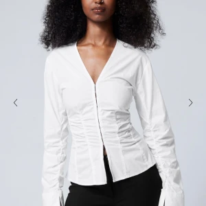 Vit blus med hakknäppning - Elegant vit blus med hakknäppning framtill och rynkade detaljer. Den har en v-ringad design och långa ärmar, perfekt för en stilren look. Lappen är kvar, oanvänd och kvar i sin påse. 