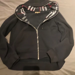 Svart Burberry - Säljer en stilren svart hoodie från Burberry med dragkedja och randigt mönster i luvan. Perfekt för en avslappnad look med en touch av lyx. Den har långa ärmar och en klassisk passform.