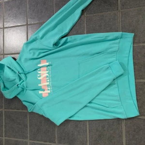 Turkos hoodie med tryck - Snygg turkos hoodie med tryck i vitt och rosa på framsidan. Den har en klassisk känguruficka och långa ärmar. Perfekt för en avslappnad stil. Storlek S men väldigt oversized så blir nog en M/L.