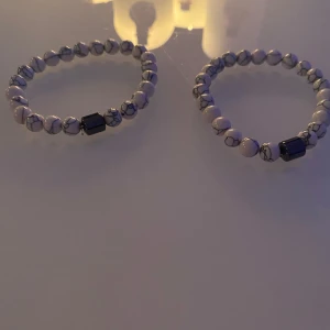 Två Vita armband med marmormönster - Två stilrena armband med vita pärlor i marmormönster och en svart rektangulär pärla som detalj. Perfekta för att ge en elegant touch till din outfit.