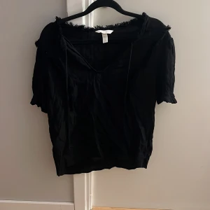 Svart topp med fransdetaljer - Snygg svart topp från H&M med fransdetaljer vid halsringningen och ärmslut. Toppen har en v-ringad design och korta ärmar, perfekt för en stilren look.