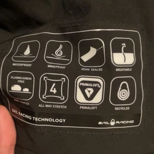 Svart jacka från Sail Racing - Säljer en svart jacka från Sail Racing med huva och dragkedja. Jackan är vattentät, vindtät och har andningsförmåga. Den är tillverkad med Primaloft och är återvunnen. Perfekt för utomhusaktiviteter. Jackan är även nästan helt ny med 