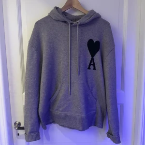 Grå hoodie från Ami Alexandre Mattiussi - Säljer en stilren grå hoodie från Ami med en svart hjärta och bokstaven 'A' på bröstet. Tröjan har en klassisk och justerbar huva med dragsko. Perfekt för en avslappnad outfit.