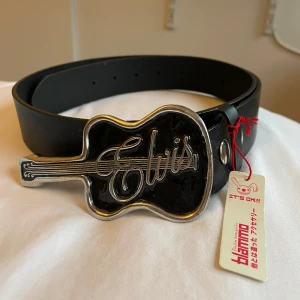 Svart bälte med Elvis-spänne - Coolt svart bälte från Blammo med ett unikt gitarrformat spänne där det står 'Elvis'. Perfekt för att ge din outfit en rockig touch. Bältet har justerbara hål för att passa olika storlekar.