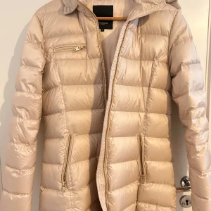 Vinterjacka från Rockandblue - Beige dunjacka från Rockandblue med huva (pälskrage finns) och dragkedjor i guld. Jackan har en quiltad design och flera praktiska fickor med dragkedja. Nyskick, endast använd en gång.  