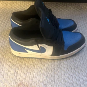Nike Air Jordan 1 Low i blå och svart - Säljer ett par Nike Air Jordan 1 Low i en snygg kombination av blått, svart och vitt. Skorna har den klassiska snörningen och den ikoniska Nike-loggan på sidan. Perfekta för dig som vill ha en stilren och sportig look. Kan gå ner lite i pris vid snabb affär.