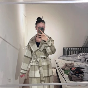 Rutig kappa från NA-KD - Säljer en stilren rutig kappa från NA-KD i beige och vit. Kappan har ett bälte i midjan och långa ärmar. Perfekt för att hålla stilen under kyligare dagar.