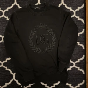 Svart Dolce & Gabbana sweatshirt - Snygg svart sweatshirt från Dolce & Gabbana med broderad logotyp och kransdetalj på framsidan. Tröjan har långa ärmar och en klassisk rund halsringning. Säljer då den blev för liten men den är riktigt fet!