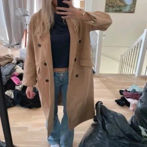 Beige kappa med knappar - Säljer en stilren beige kappa med knappar framtill. Den har en klassisk design med långa ärmar och två fickor. Perfekt för höst och vår.