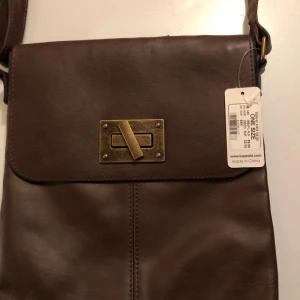 Brun axelväska med metallspänne - Snygg brun axelväska i syntetmaterial med ett stilrent metallspänne framtill. Väskan har en justerbar axelrem och en praktisk storlek för vardagsbruk. Perfekt för att bära med sig det nödvändigaste.