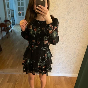 Klänning med volanger💗 - Jättefin svart klänning med blommigt mönster i rosa och grönt. Klänningen har långa ärmar och volangdetaljer i kjolen. Perfekt för en stilfull look. Den säger barnstorlek 158 men passar som xs/s💗💗 Den har inga defekter eller tecken på användning🥰