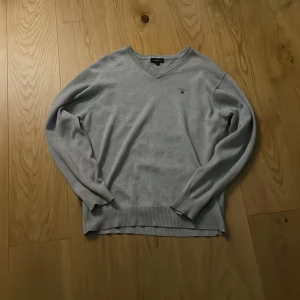 Grå tröja från GANT - Säljer en stilren grå tröja från GANT med v-ringning och broderad logotyp på bröstet. Tröjan har långa ärmar och ribbstickade muddar vid ärmslut och nederkant. Perfekt för en avslappnad och snygg look.
