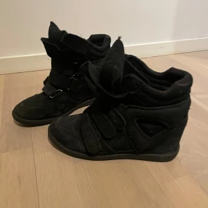 Trendiga svarta sneakers med kilklack - Snygga svarta sneakers med kilklack och kardborreband. Skorna har en unik design med högre skaft och är tillverkade i mocka. Perfekta för en trendig look. Stl 40. 