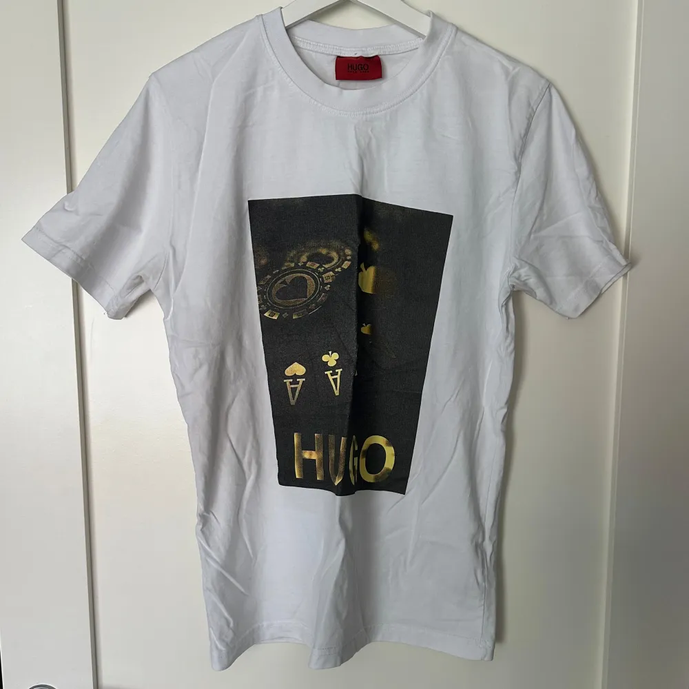 Säljer en snygg vit t-shirt från Hugo Boss i storlek M. Den har ett coolt tryck i svart och guld med spelkort och texten 'HUGO' på framsidan. Perfekt för en avslappnad stil eller en utekväll. T-shirten är i bomull och har korta ärmar.skick 9/10. Info. T-shirts.