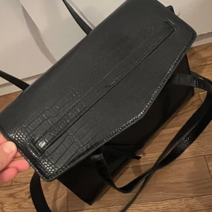 Svart axelväska i syntet med krokodilmönster - Säljer en snygg svart axelväska med krokodilmönster. Väskan är gjord i syntetmaterial och har en stilren design med ett lock som stängs med en knapp. Perfekt för både vardag och fest!