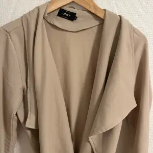 Säljer en snygg beige trenchcoat från ONLY. Den har en stilren design med ett knytbälte i midjan och långa ärmar. Perfekt för vår och höst!