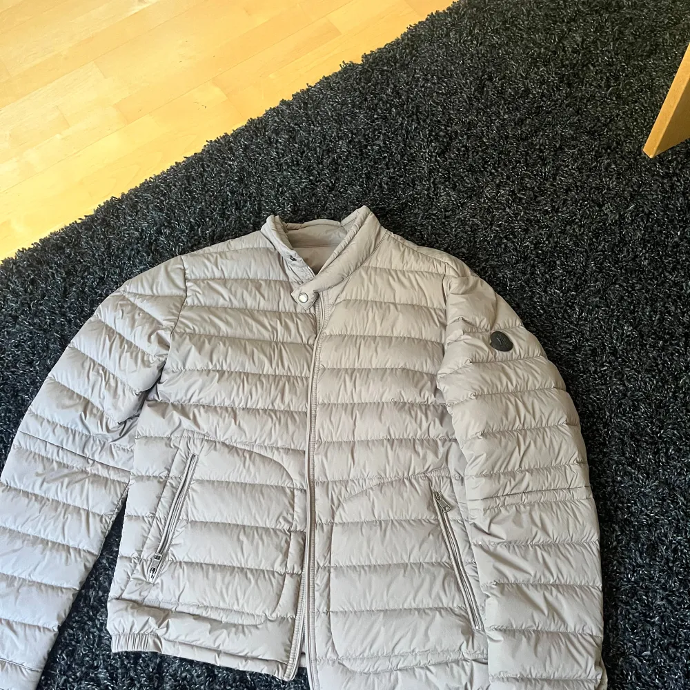 Säljer en snygg och stilren grå dunjacka från Moncler. Jackan är quiltad och har en dragkedja framtill. Den är perfekt för kyligare dagar och har en normal passform. Moncler-loggan syns tydligt på ärmen, vilket ger en exklusiv touch.. Jackor.