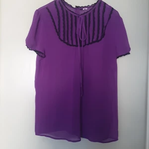 Lila blus med svarta detaljer - Säljer en lila blus med svarta detaljer och knappar på baksidan. Blusen har korta ärmar och en dekorativ knytning framtill. Perfekt för både vardag och fest!