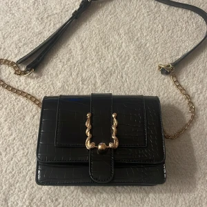 Svart axelväska med guldkedja - Säljer en superfin svart axelväska i syntetmaterial med krokodilpräglat mönster. Väskan har en elegant guldkedja och en snygg detalj framtill. Perfekt för både vardag och fest!