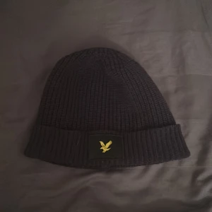 Svart ribbad mössa Lyle Scott - Snygg svart ribbad mössa med en broderad gul logga framtill. Perfekt för att hålla värmen under kyliga dagar. Enkel och stilren design som passar till det mesta.