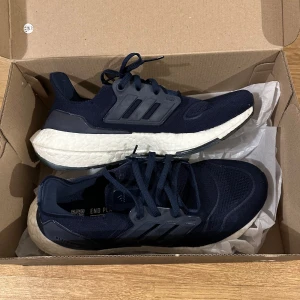Adidas Ultraboost 22 Navy/Svart  - Säljer ett par svarta Adidas Ultraboost med vit sula. Skorna har en stilren design med snörning och är perfekta för både träning och vardag. De är tillverkade i syntetmaterial för optimal komfort och hållbarhet. Nyskick och endast provade. Nypris 2049 kr. 