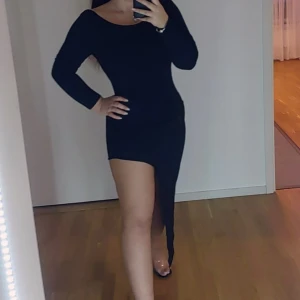 Svart klänning med öppen rygg - Snygg svart klänning med lång ärm och en djärv öppen rygg. Den har en asymmetrisk slits som ger en extra touch av stil. Perfekt för en kväll ute!