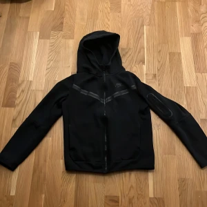 Svart jacka med huva från Nike - Snygg svart jacka från Nike med huva och dragkedja framtill. Jackan har en diskret logga på bröstet och en praktisk ficka med dragkedja på ärmen. Perfekt för en sportig look.