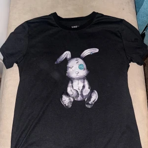 Svart t-shirt med kaninmotiv från Shein - Snygg svart t-shirt från Shein med ett unikt kaninmotiv på framsidan. Tillverkad i en mjuk blandning av bomull och polyester. Perfekt för en avslappnad stil.