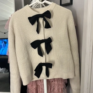 Vit stickad kofta med svarta rosetter - Säljer en supersöt vit stickad kofta med tre svarta rosetter framtill. Koftan är endast använd en gång så i princip helt ny🥰 