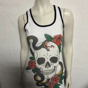Linne från Insanity M/L - Coolt vitt linne med ett stort tryck av en dödskalle, ormar och rosor. Linnet har svarta kanter vid ärmhål och halsringning. Perfekt för en edgy look!