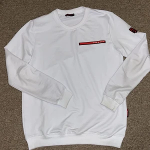 Vit sweatshirt - Säljer en stilren vit tröja från Prada med en röd logga på bröstet. Tröjan har långa ärmar och ribbade muddar vid ärmslut och nederkant. Perfekt för en minimalistisk look. Inga skador eller nåt sånt, kan mötas upp i stockholm men även frakta!  