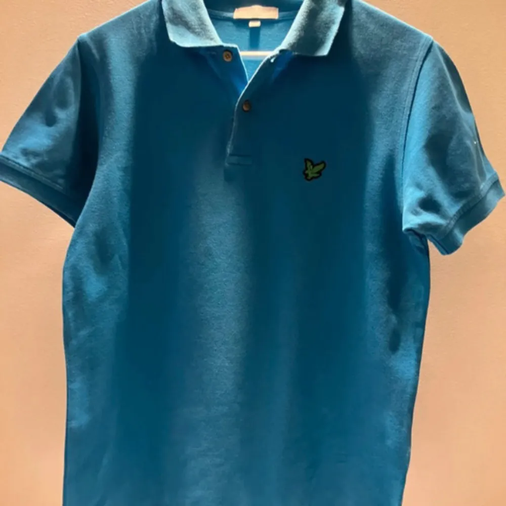 Snygg blå pikétröja från Lyle & Scott med klassisk krage och knappslå. Tröjan har en broderad logotyp på bröstet och korta ärmar. Perfekt för en stilren look.. T-paidat.