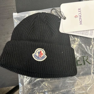 Svart mössa från Moncler - Snygg svart ribbad mössa från Moncler med broderad logotyp framtill. Perfekt för att hålla värmen under kyliga dagar. Mössan har en klassisk design som passar till det mesta.