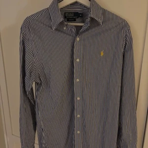 Randig skjorta från Ralph Lauren - Snygg blå- och vitrandig skjorta från Ralph Lauren i slim fit. Skjortan har långa ärmar och klassisk krage med knappar framtill. Den ikoniska loggan är broderad på bröstet. Perfekt för en stilren look!