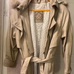 Beige läderkappa med pälsdetaljer XS/S - Snygg beige läderkappa med pälsdetaljer på huvan och quiltat foder. Jackan har ett bälte i midjan och dragkedja framtill. Perfekt för kyligare dagar med sin långa design och stilrena look.