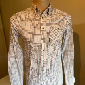 Rutig skjorta från Brokared Tattersallskjorta - Snygg Tattersallskjorta rutig skjorta från Brokared i vitt med blå och röda rutor. Skjortan har långa ärmar och en klassisk krage. Den är försedd med knappar och en bröstficka för extra stil och funktionalitet. Brittisk jaktstil 