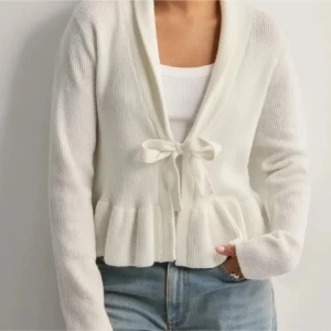 Vit stickad kofta med knytband - Säljer en söt vit stickad kofta med knytband framtill. Koftan har långa ärmar och en volangdetalj vid nederkanten. Perfekt för en mysig och stilren look. Från Nelly och endast använd en gång! Jätte mysig❤️ hör av er vid frågor, jag postar så snart jag kan!