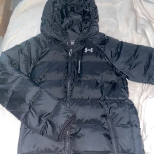 Svart pufferjacka från Under Armour - Säljer en svart pufferjacka från Under Armour med huva och dragkedja. Jackan har ett stilrent och sportigt utseende med märkets logga på bröstet. Perfekt för kyligare dagar. Den kommer ej till användning så säljer den köpte den för 1299 o säljer den för endast 1099