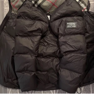 Svart dunväst från Burberry - Snygg svart dunväst från Burberry med klassiskt rutigt mönster på insidan. 