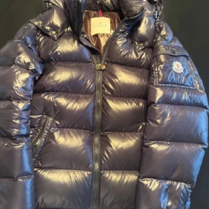 Svart dunjacka från Moncler - Snygg och varm svart dunjacka från Moncler med glansig finish. Jackan har en dragkedja framtill och en broderad Moncler-logga på ärmen. Perfekt för kyliga dagar.