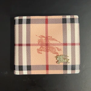  plånbok från Burberry - Snygg rutig plånbok från Burberry i beige och bruna toner med klassiskt mönster. Plånboken har flera kortfack och en genomskinlig ID-ficka. Tillverkad i skinn med en elegant guldlogga på framsidan. Perfekt för att hålla ordning på dina kort och kontanter.