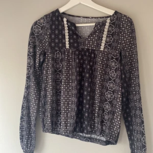 Mönstrad långärmad blus - Säljer en unik mönstrad blus i svart och grått med blommönster och spetsdetaljer. Blusen har en v-ringad hals och långa ärmar, perfekt för en stilfull look.